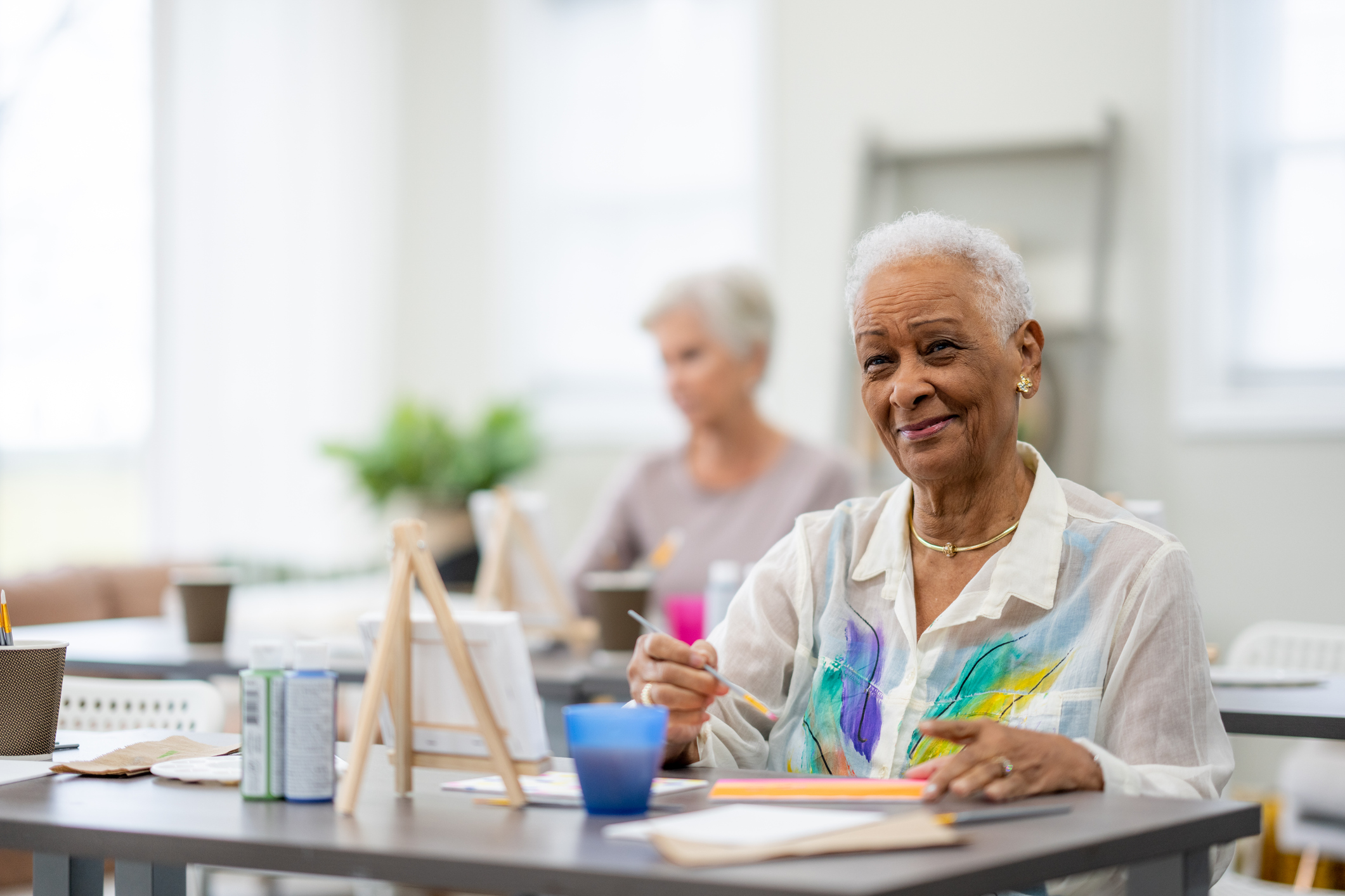 A small group of senior citizens sit together in an art class as they work on individual paintings.  The focus is on a woman of African decent in front who is diligently painting away.